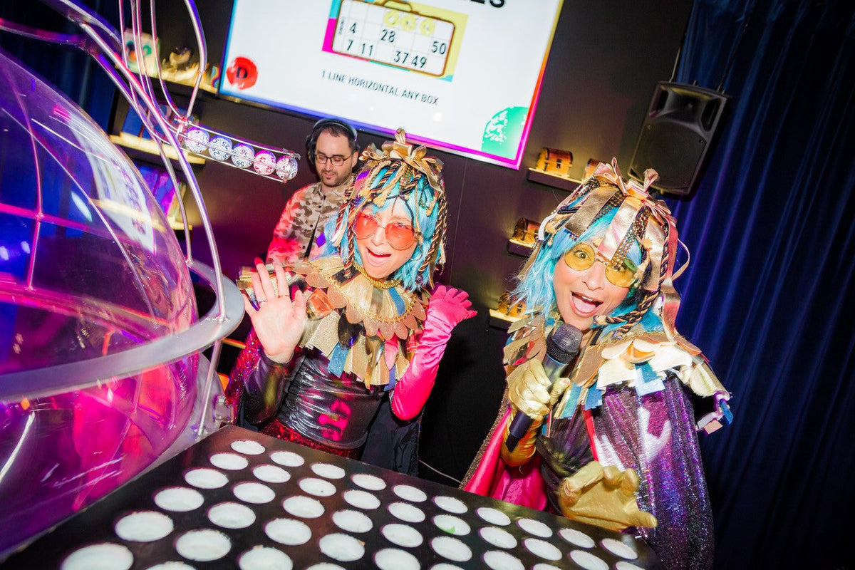 Two vibrant performers in colorful costumes and wigs engage with the audience at an interactive event.